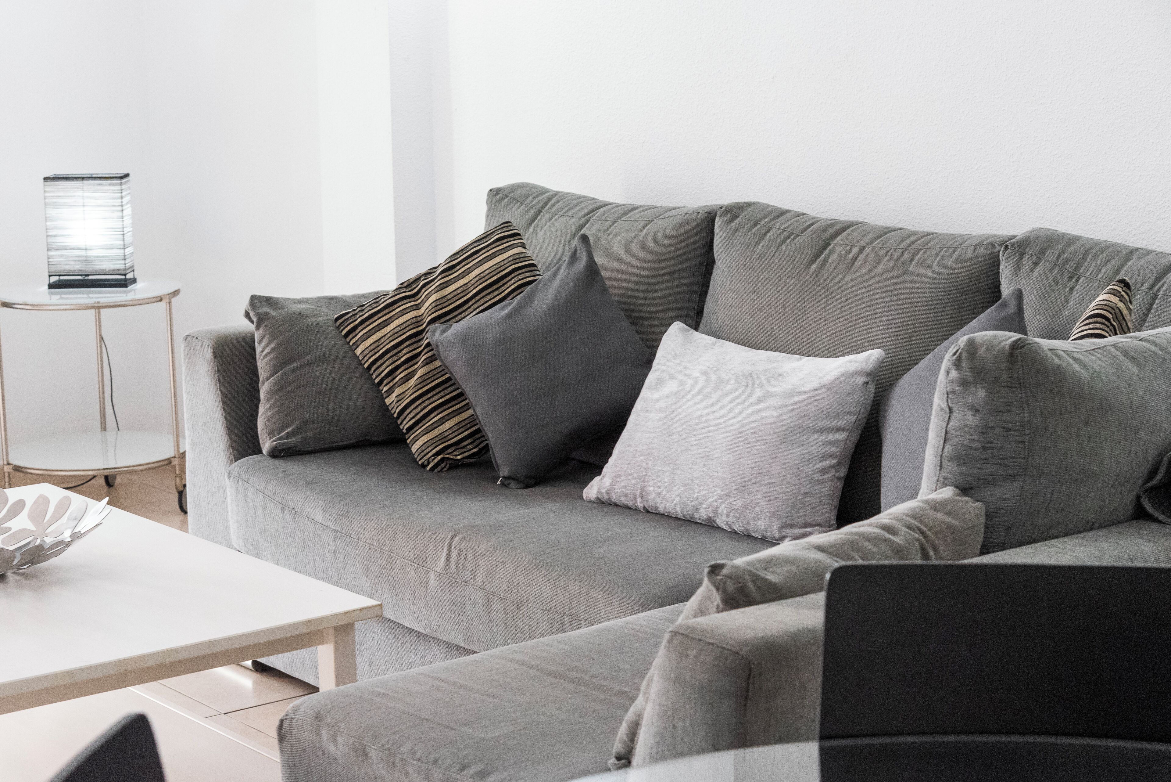 Ein graues Sofa mit mehreren Kissen, ein weißer Beistelltisch und ein kleiner runder Tisch mit einer Lampe.