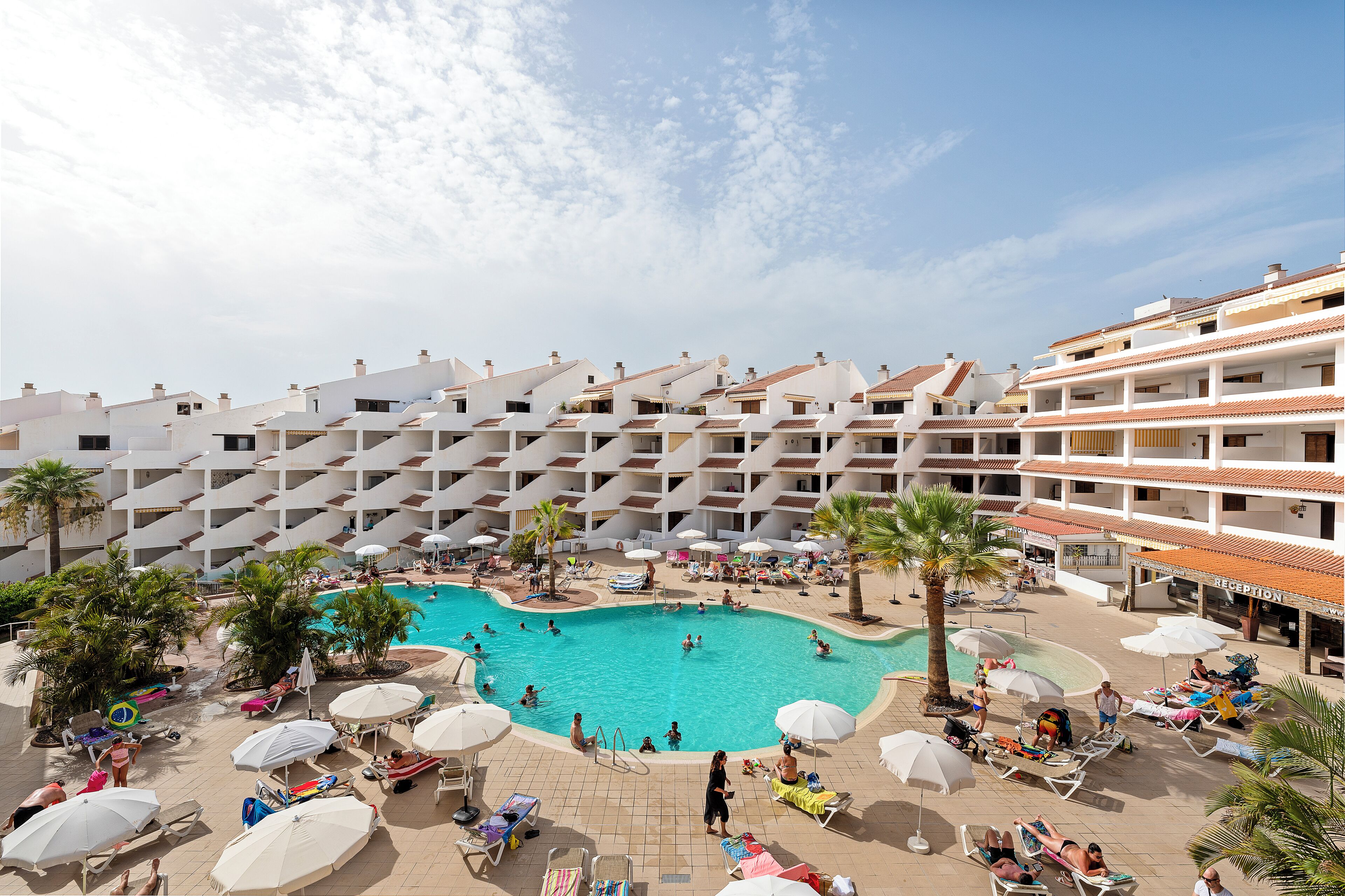 A resort with a central swimming pool surrounded by sun loungers and umbrellas, a reception area, and multiple balconies.