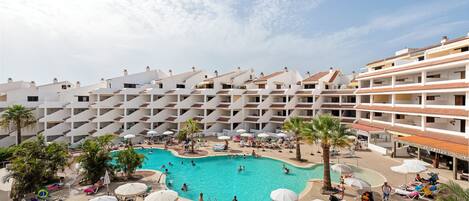 A resort with a central swimming pool surrounded by sun loungers and umbrellas, a reception area, and multiple balconies.