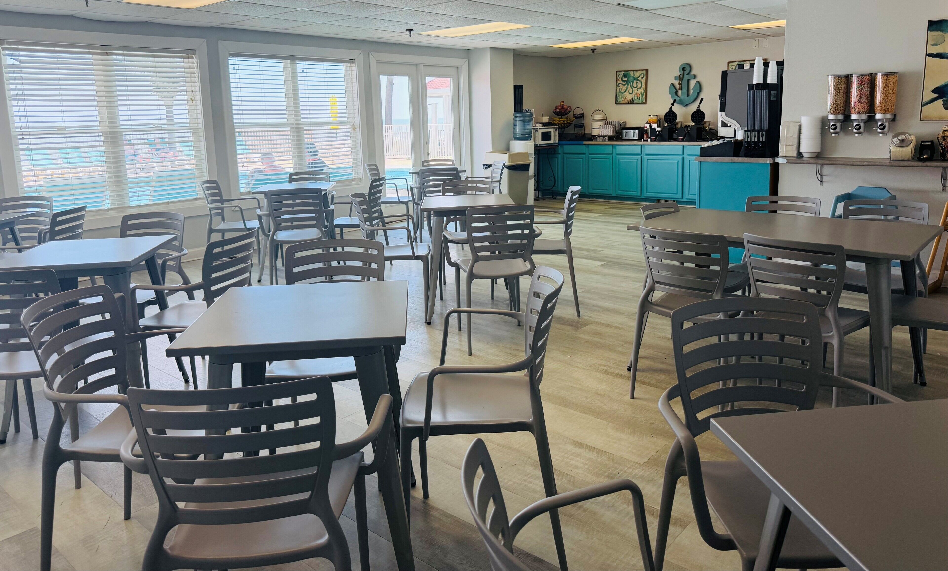 A cafeteria with tables and chairs, a kitchen area, and a view through large windows.
