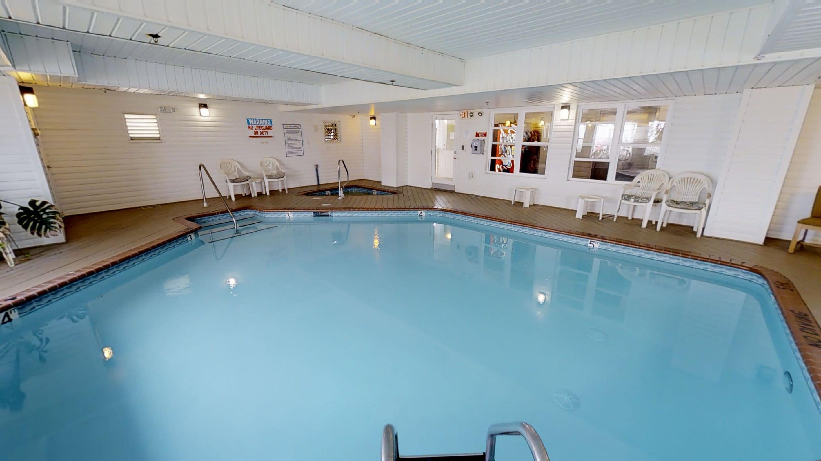 An indoor swimming pool with a wooden deck, white walls, and a lifeguard warning sign.