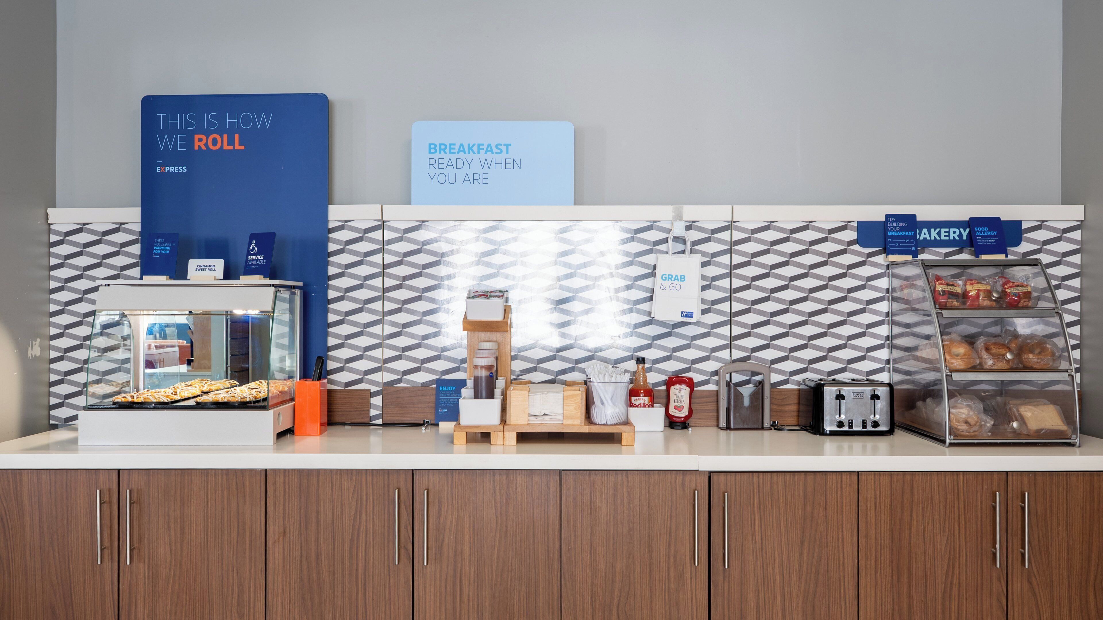 A breakfast counter with a sign indicating 'Breakfast Ready When You Are,' a display case with pastries, a sweet roll service sign, and a grab & go sign.