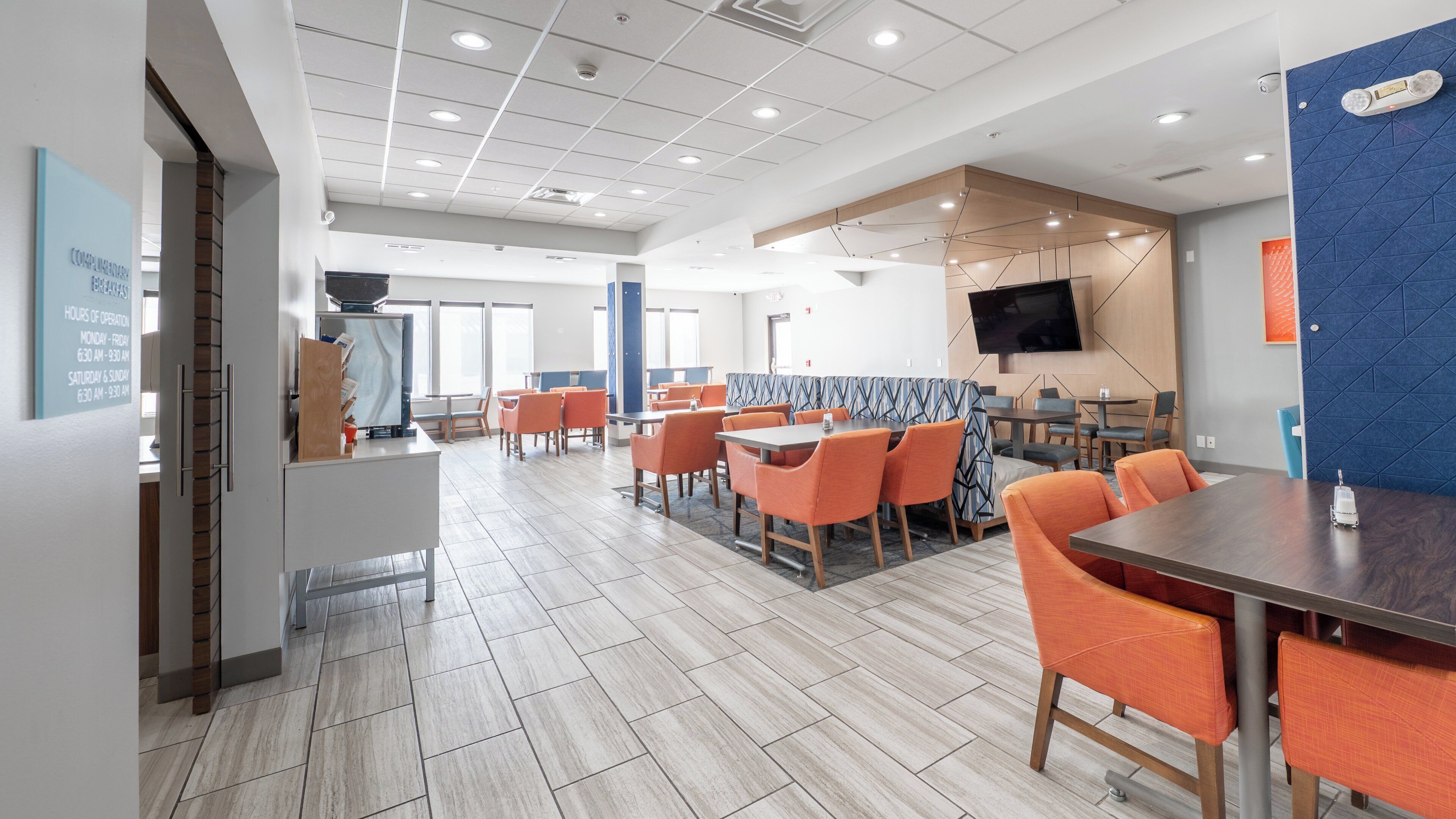 A spacious dining area with multiple tables and chairs, a flat-screen TV mounted on the wall, and a sign indicating hours of operation.