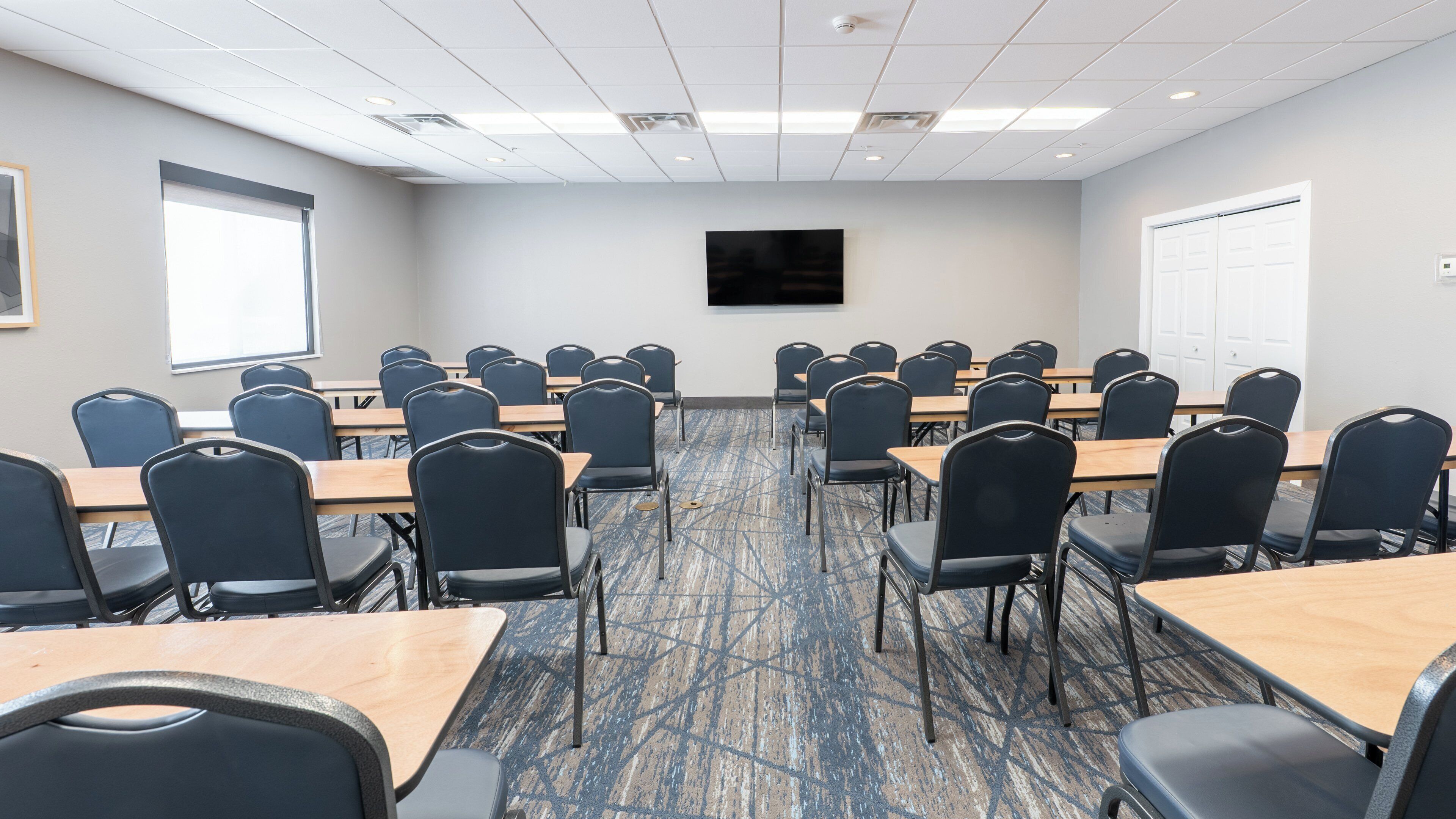 A conference room with rows of tables and chairs, a flat-screen TV on the wall, and a door on the right.