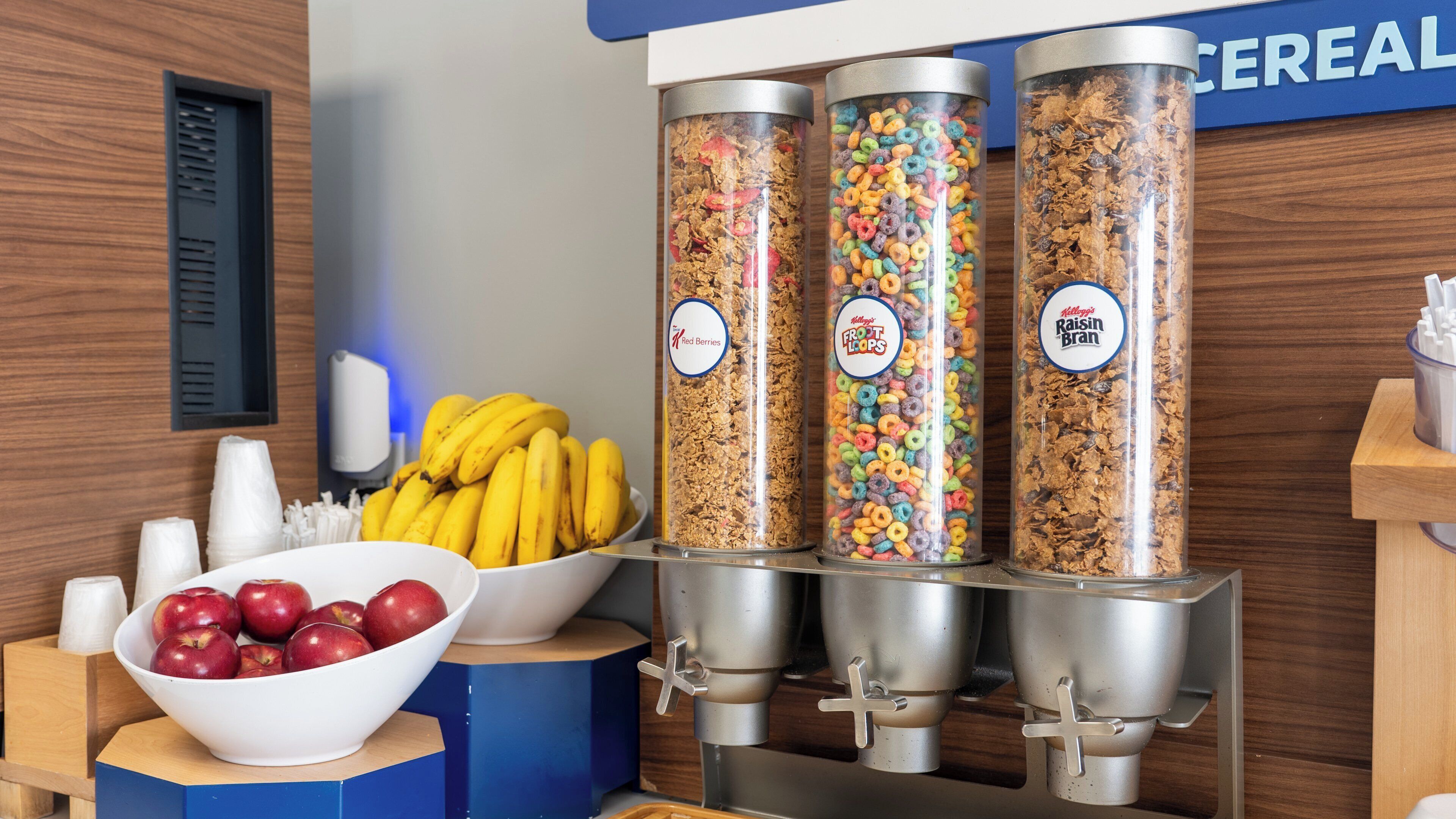 En morgenmadsbar med tre skåle til cornflakes, mærket 'Red Berries', 'Frost Loops' og 'Raisin Bran'. Skåle med æbler og bananer, og et blåt skilt i baggrunden.