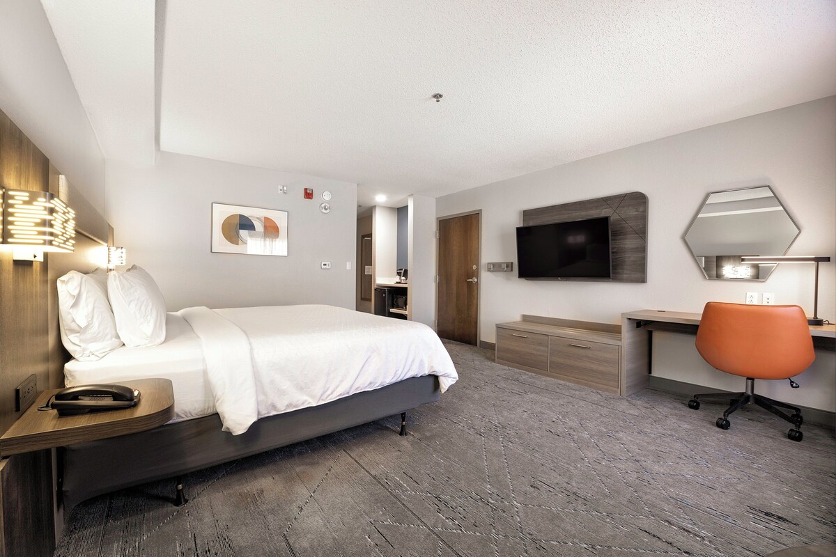 A modern hotel room with a bed, bedside table, desk, chair, and a flat-screen TV mounted on the wall.