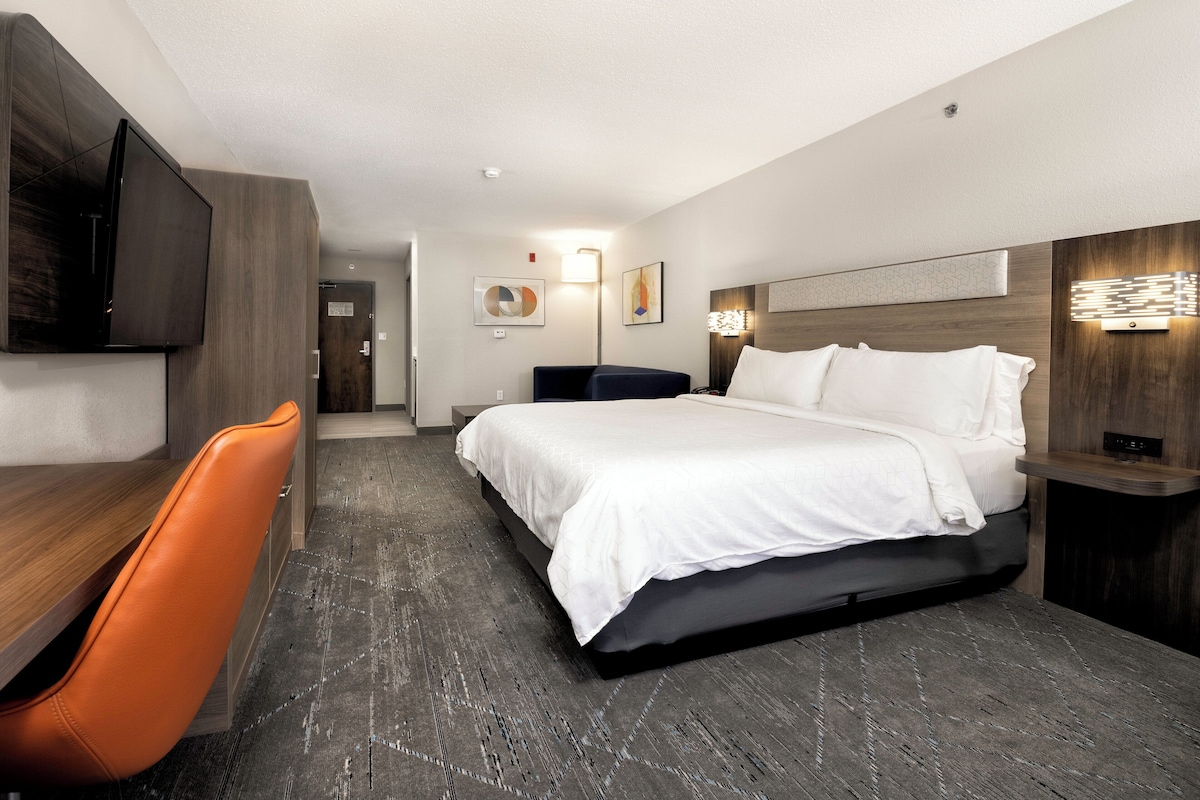 A modern hotel room with a large bed, a desk with a chair, a flat-screen TV, and a wall-mounted light fixture.