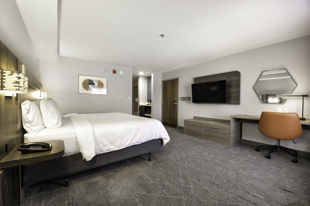 A modern hotel room with a bed, bedside table, desk, chair, and a flat-screen TV mounted on the wall.