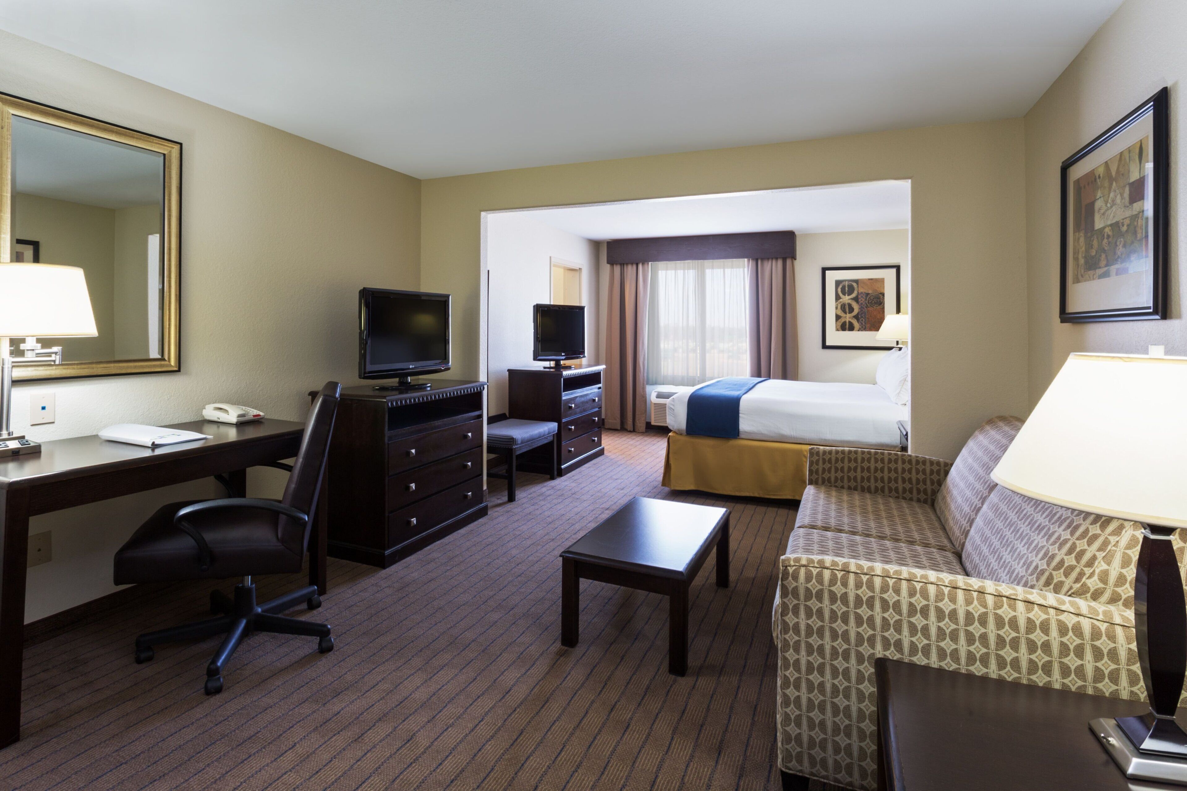 A hotel room with a desk, chair, television, bed, sofa, and a large mirror.