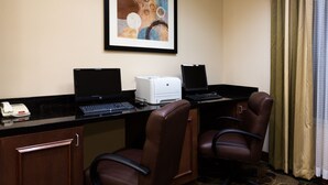 A hotel room with two desks, each with a laptop, a printer, and a telephone. There are two chairs and a door.