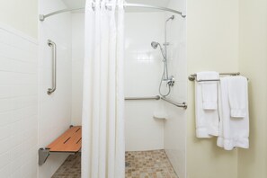 Un baño con paredes de azulejos blancos, una ducha con puerta de cristal, una barra de apoyo, un banco y un perchero con toallas.