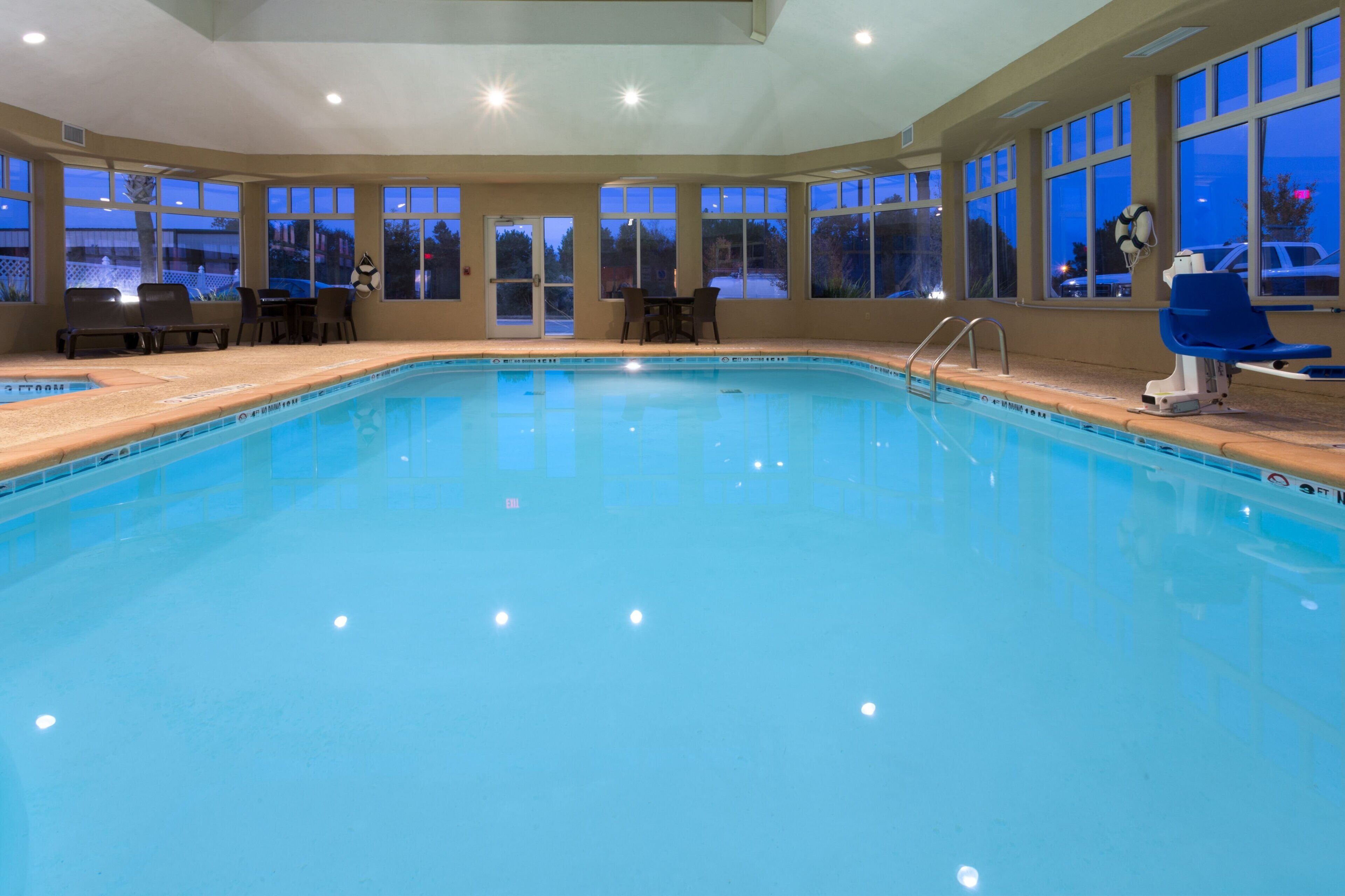 An indoor swimming pool with clear blue water, surrounded by windows and chairs.