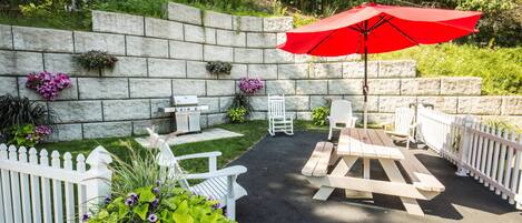 Un patio con muro de piedra, cerca de piquetes blancos y mobiliario exterior, incluyendo una mesa y sillas.
