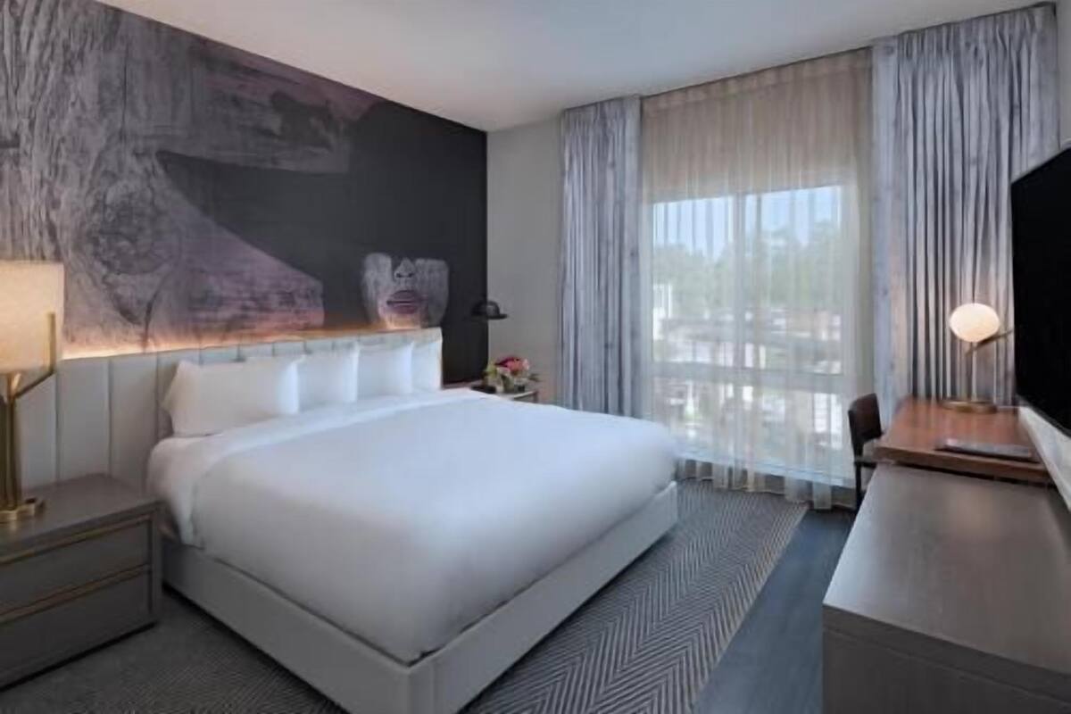 Quarto de hotel com uma cama grande, televisão de tela plana, escrivaninha com luminária e janela com cortinas.