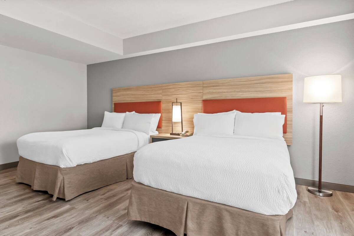 Una habitación de hotel con dos camas, cada una con ropa de cama blanca y un cabecero de madera. Hay una mesita de noche con una lámpara entre las camas.