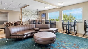 Un hall d’hôtel moderne avec un canapé d’angle en cuir marron, un pouf rond et un coin repas comprenant des chaises et des tables en bois.