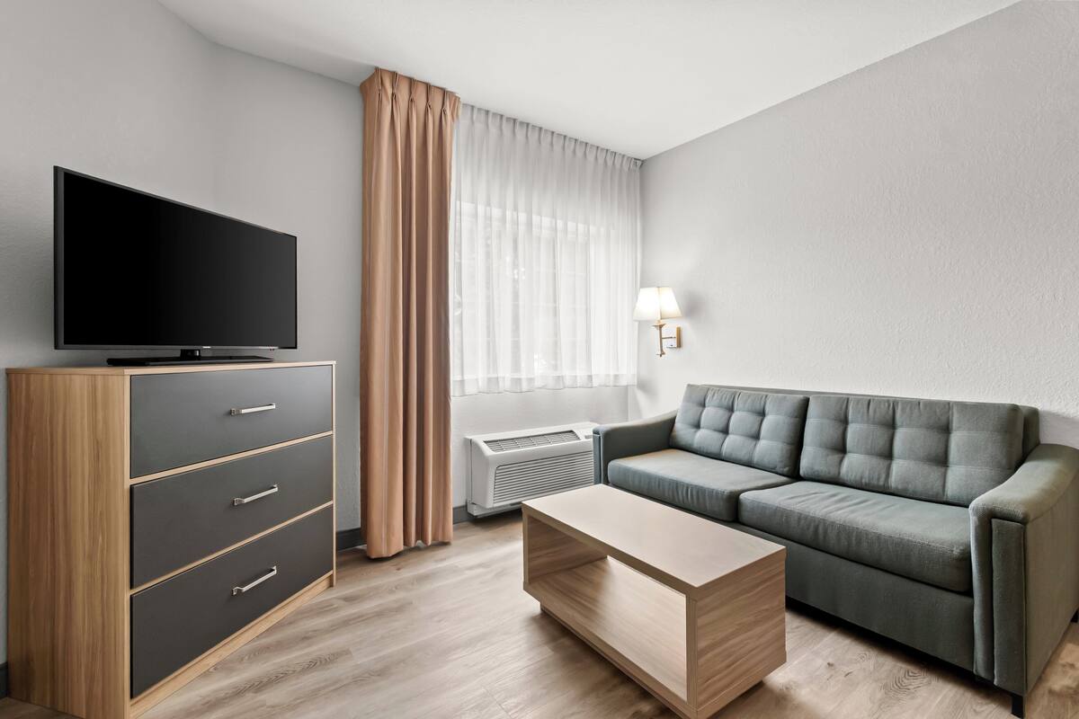 Una sala de estar moderna con un televisor de pantalla plana, un mueble de madera con cajones, un sofá gris, una mesa de centro de madera y una ventana con cortinas traslúcidas.