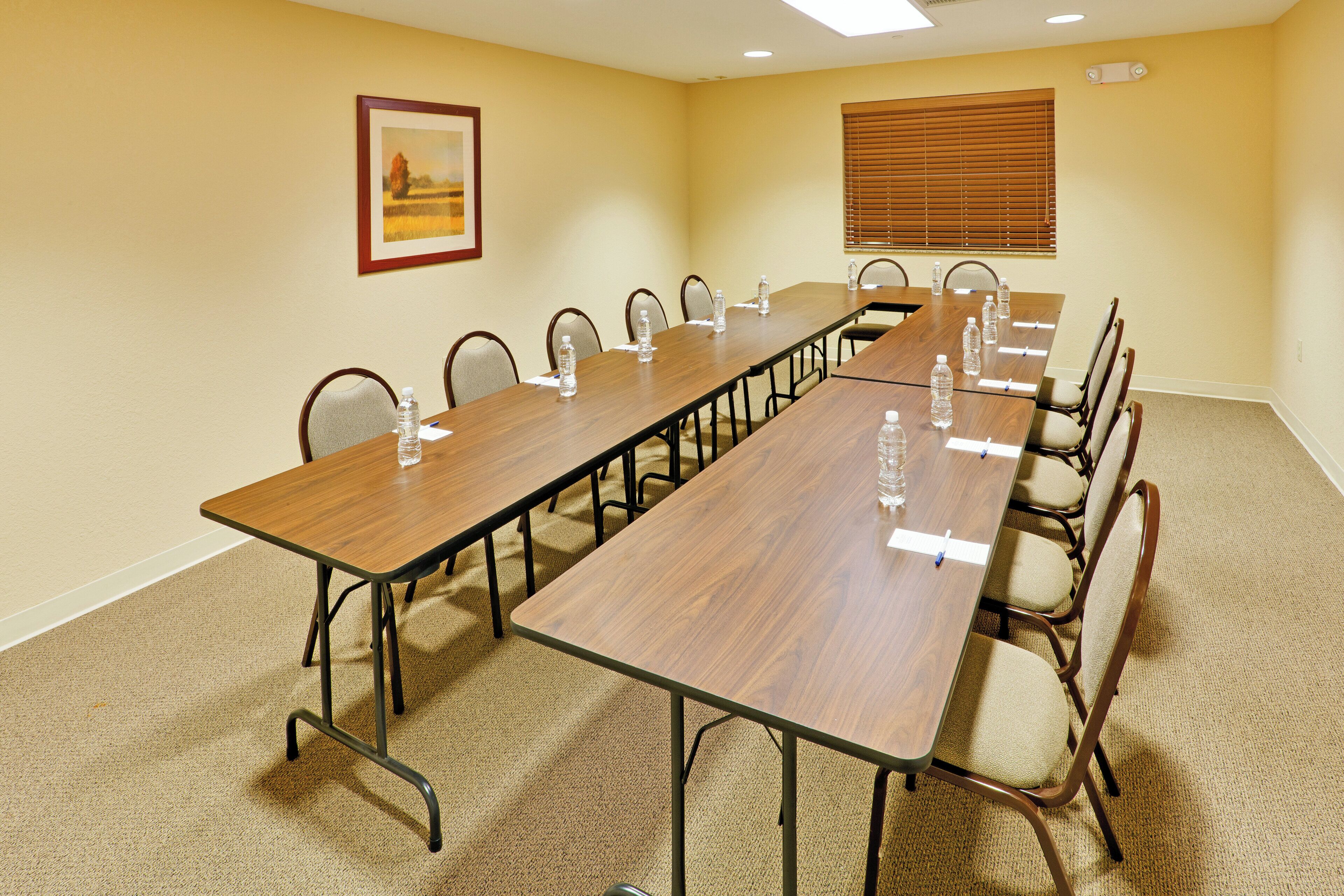 A conference room with a long table, chairs, water bottles, a painting, and a window with blinds.