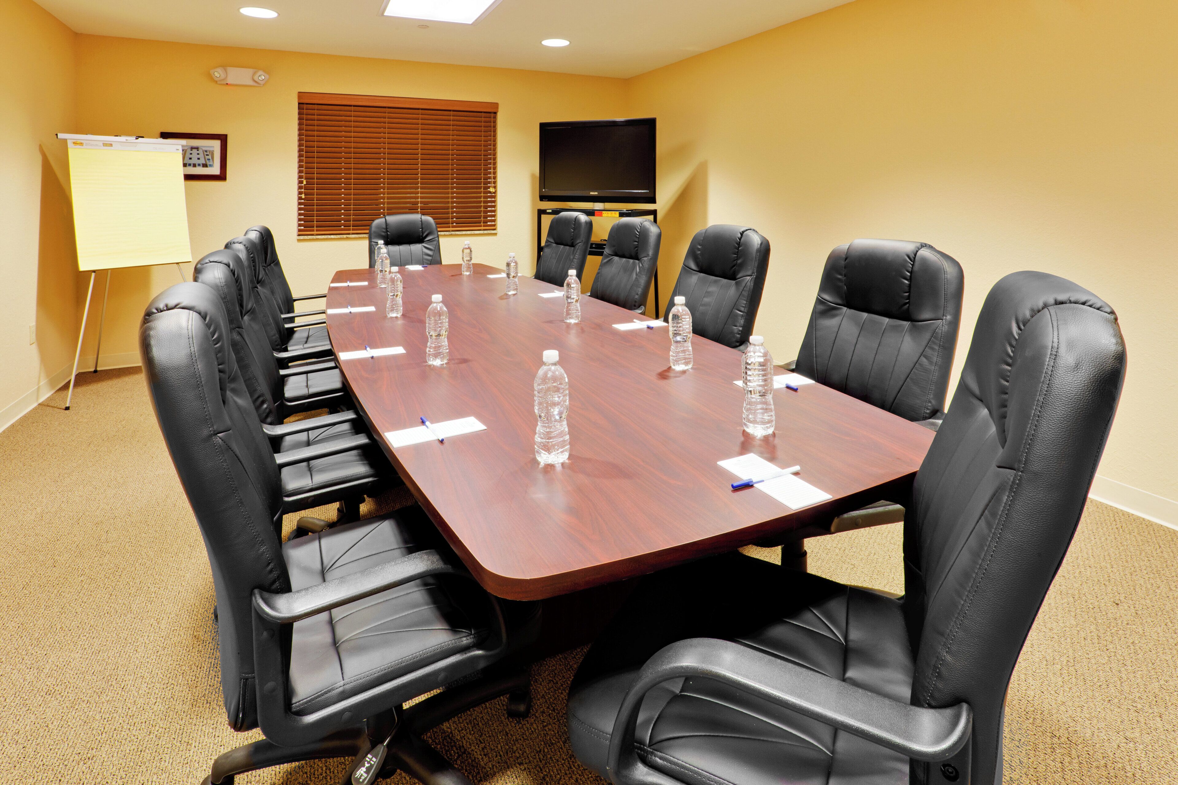 A conference room with a long table, black leather chairs, water bottles, a whiteboard, and a flat-screen TV on the wall.