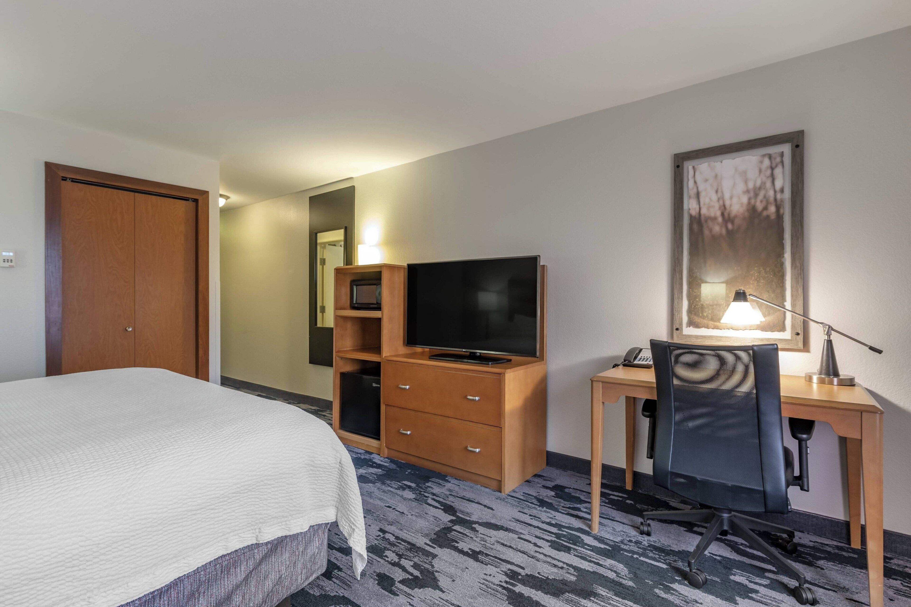 Una habitación de hotel con una cama, un escritorio de madera con una computadora, un televisor y una lámpara.