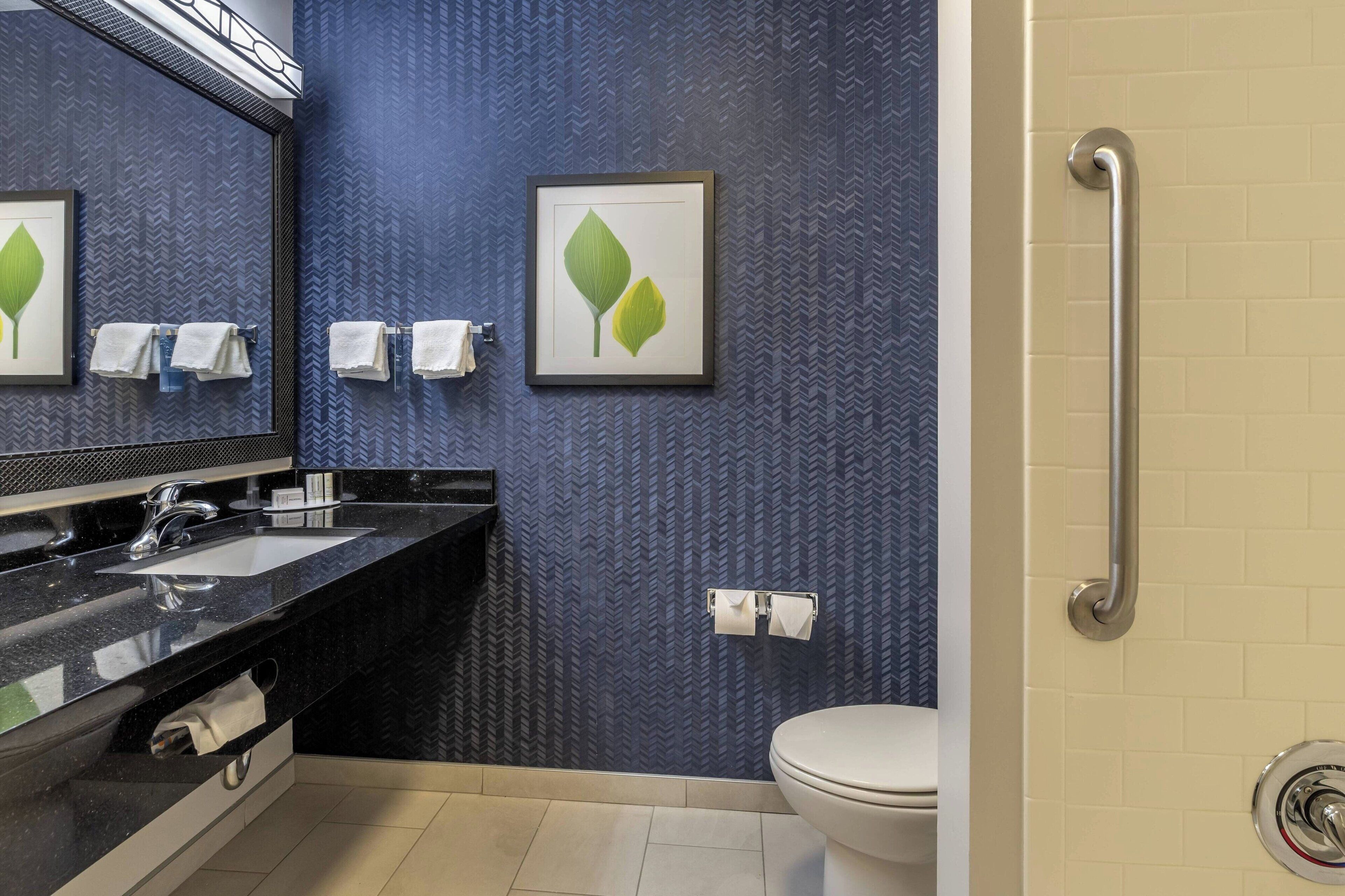 A modern bathroom with a black countertop, a sink, a mirror, and a toilet. There are towels hanging on a rack and a framed picture on the wall.
