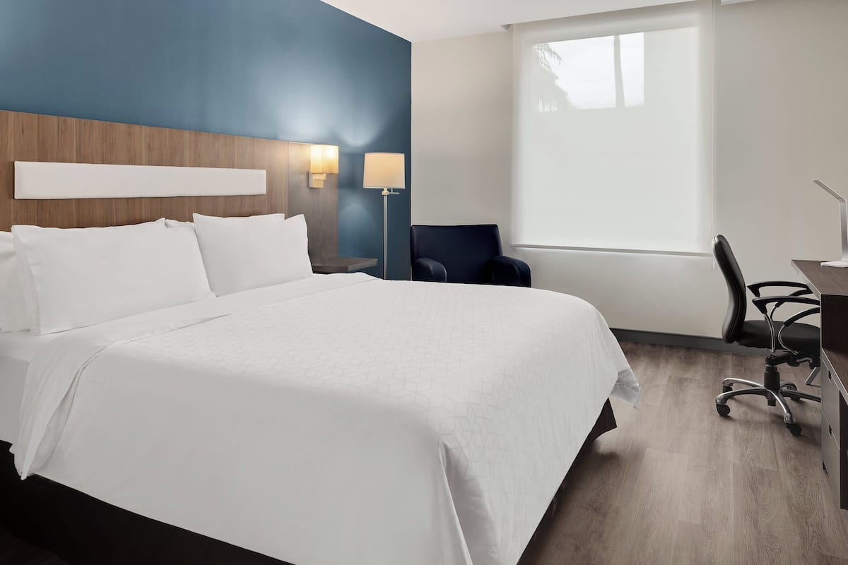 Habitación de hotel con una cama grande, un escritorio con silla, una lámpara y una ventana con persianas.
