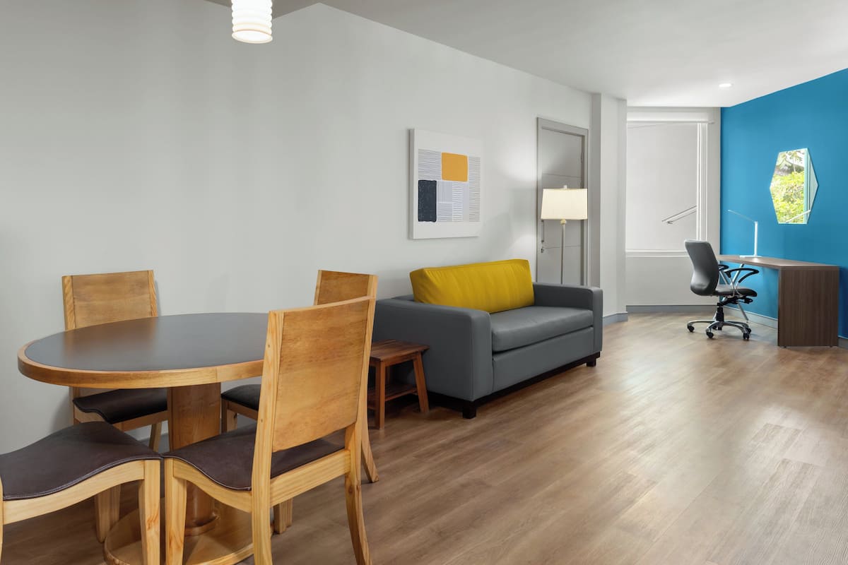 Una sala de estar moderna con una mesa redonda de madera, cuatro sillas, un sofá gris con un cojín amarillo, una pared de acento azul y un escritorio con una silla.