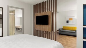 A modern hotel room with a large bed, a mounted TV, wooden paneling, a gray sofa with a yellow cushion, and a framed picture on the wall. - Holiday Inn Express San Jose Forum by IHG (Pozos)