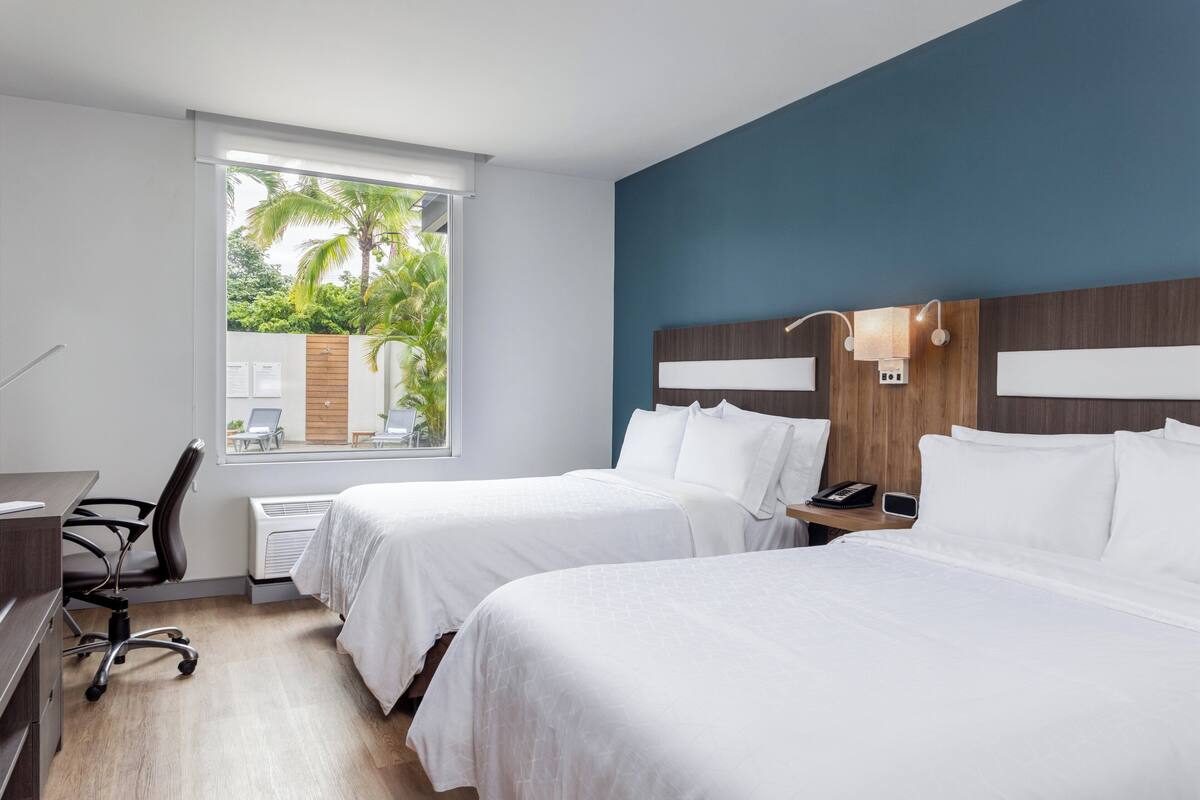 Habitación de hotel con dos camas, un escritorio, una silla y una ventana con vistas a zonas verdes.