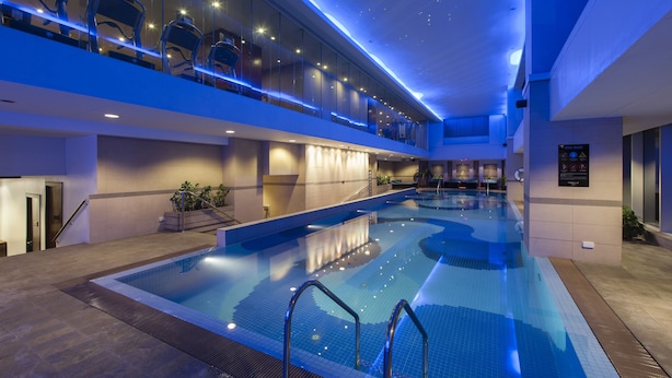 A modern indoor swimming pool with a glass ceiling, seating area, and a staircase leading to an upper level.