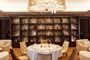 A dining area with a round table set for six, surrounded by upholstered chairs. Behind the table, there are multiple shelves displaying various decorative items and bottles. - Joy Nostalg Hotel and Suites Manila Managed by The Ascott Limited (Pasig)