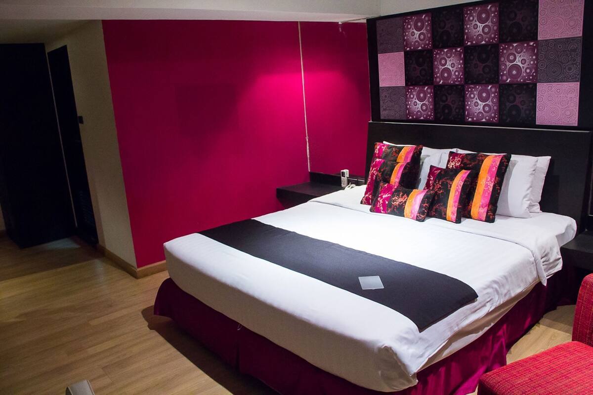 A hotel room with a bed, a red wall, a patterned wall decoration, and a wooden floor.