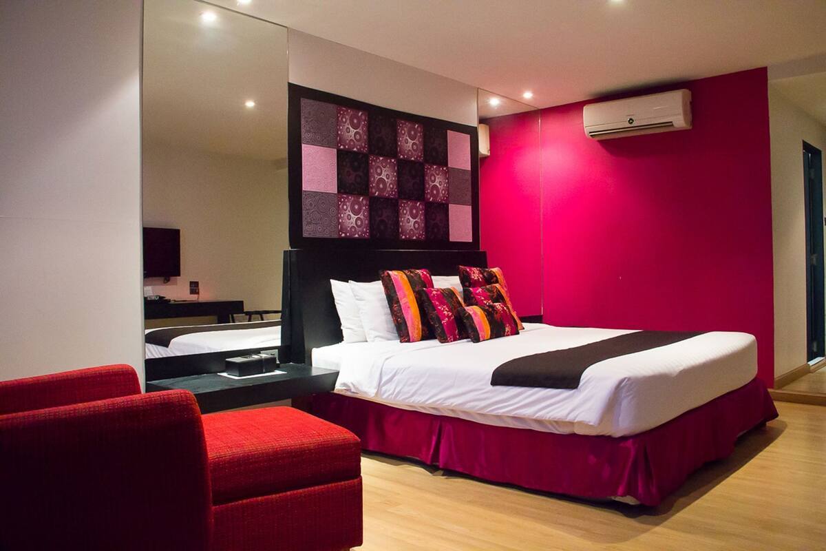 A modern hotel room with a large bed, a red armchair, a bedside table, and a wall-mounted air conditioner.