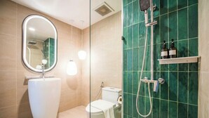 A modern bathroom with a large oval mirror, a pedestal sink, a toilet, and a shower with a handheld showerhead. - Grand Inn Hotel (Bangkok)