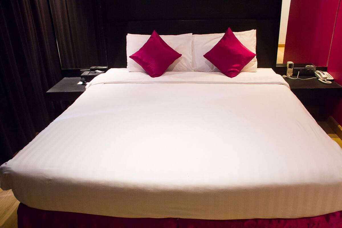 A neatly made bed with white linens and two bright pink pillows, a bedside table with a phone, and a dark-colored headboard.