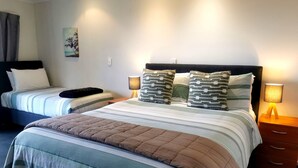 A hotel room with two beds, a large bed with a striped bedspread and pillows, and a smaller bed with a plain bedspread. There are two bedside tables with lamps, a wall art piece, and a window with curtains.