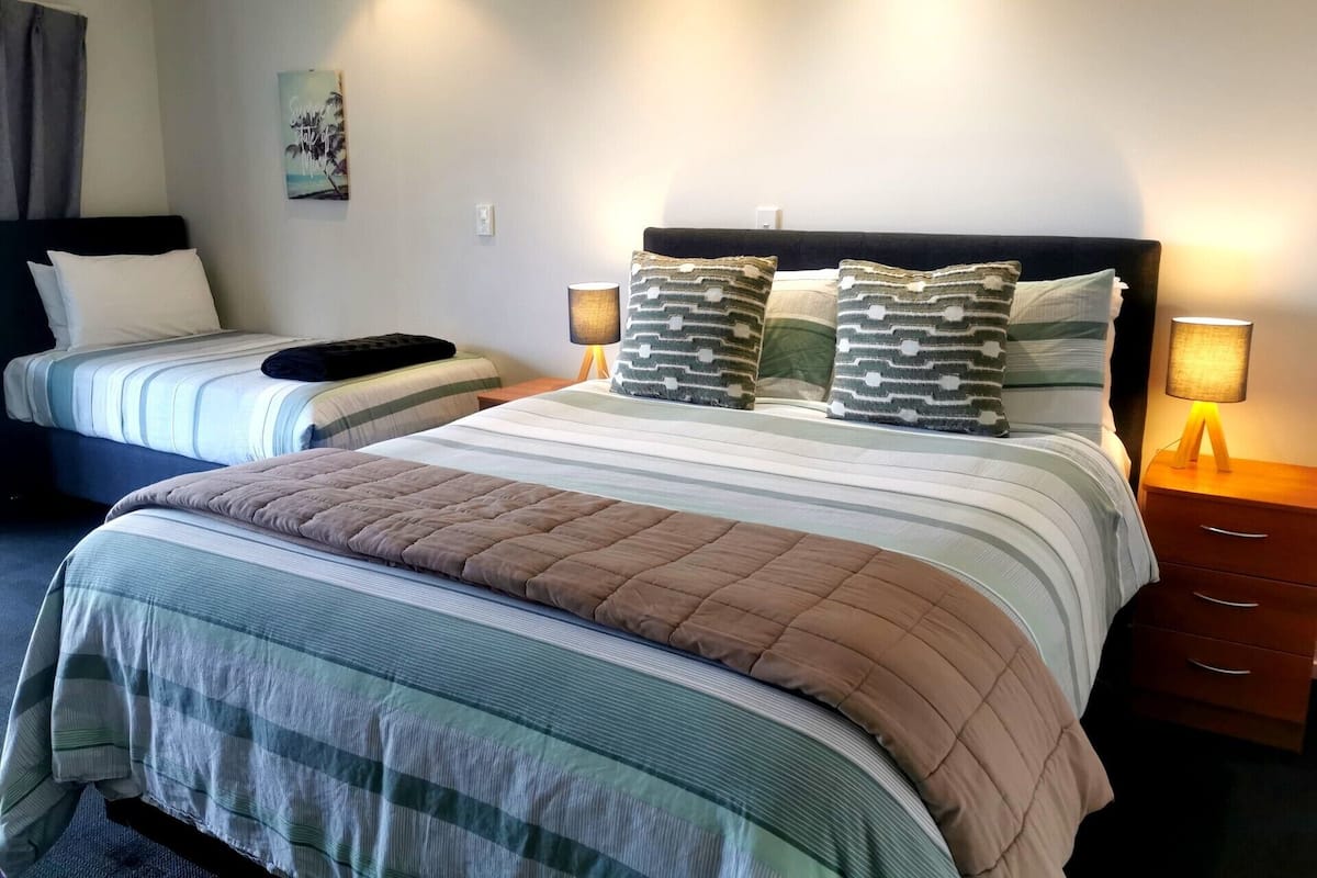 Ein Hotelzimmer mit zwei Betten, einem großen Bett mit gestreifter Bettdecke und Kissen sowie einem kleineren Bett mit schlichter Bettdecke. Zwei Nachttische mit Lampen, ein Wandbild und ein Fenster mit Vorhängen sind ebenfalls vorhanden.