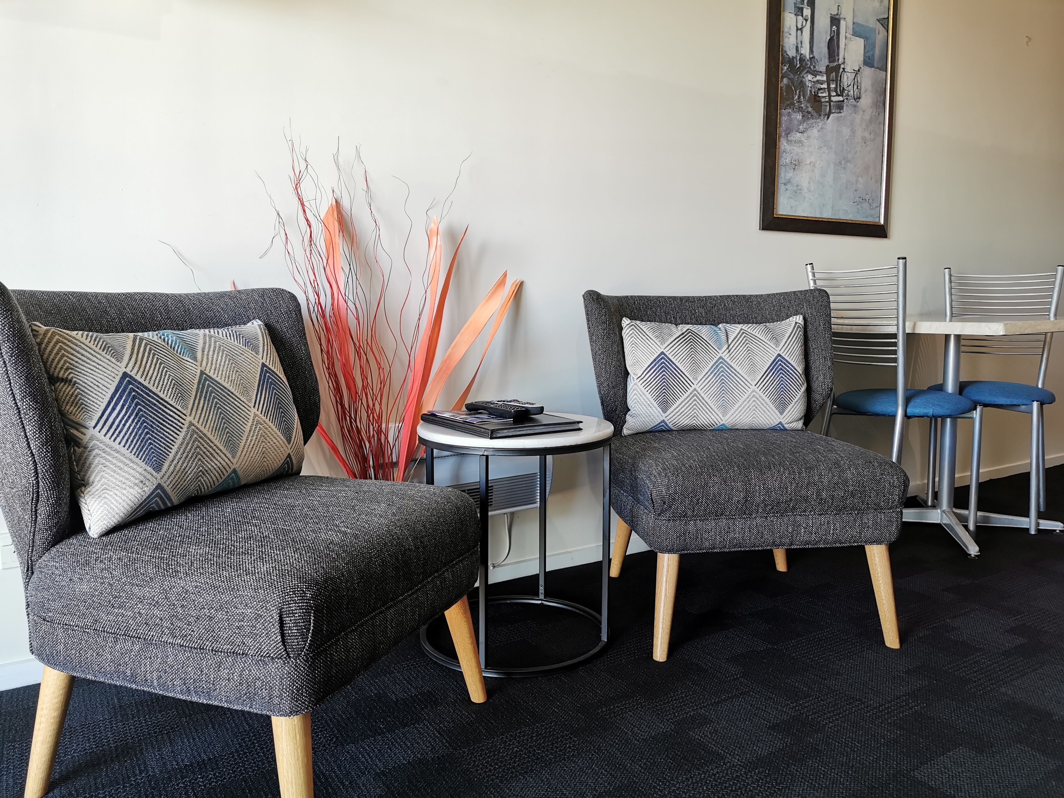 A modern room with two grey upholstered chairs with patterned cushions, a small round table with a laptop and remote, and a tall vase with red and orange branches.
