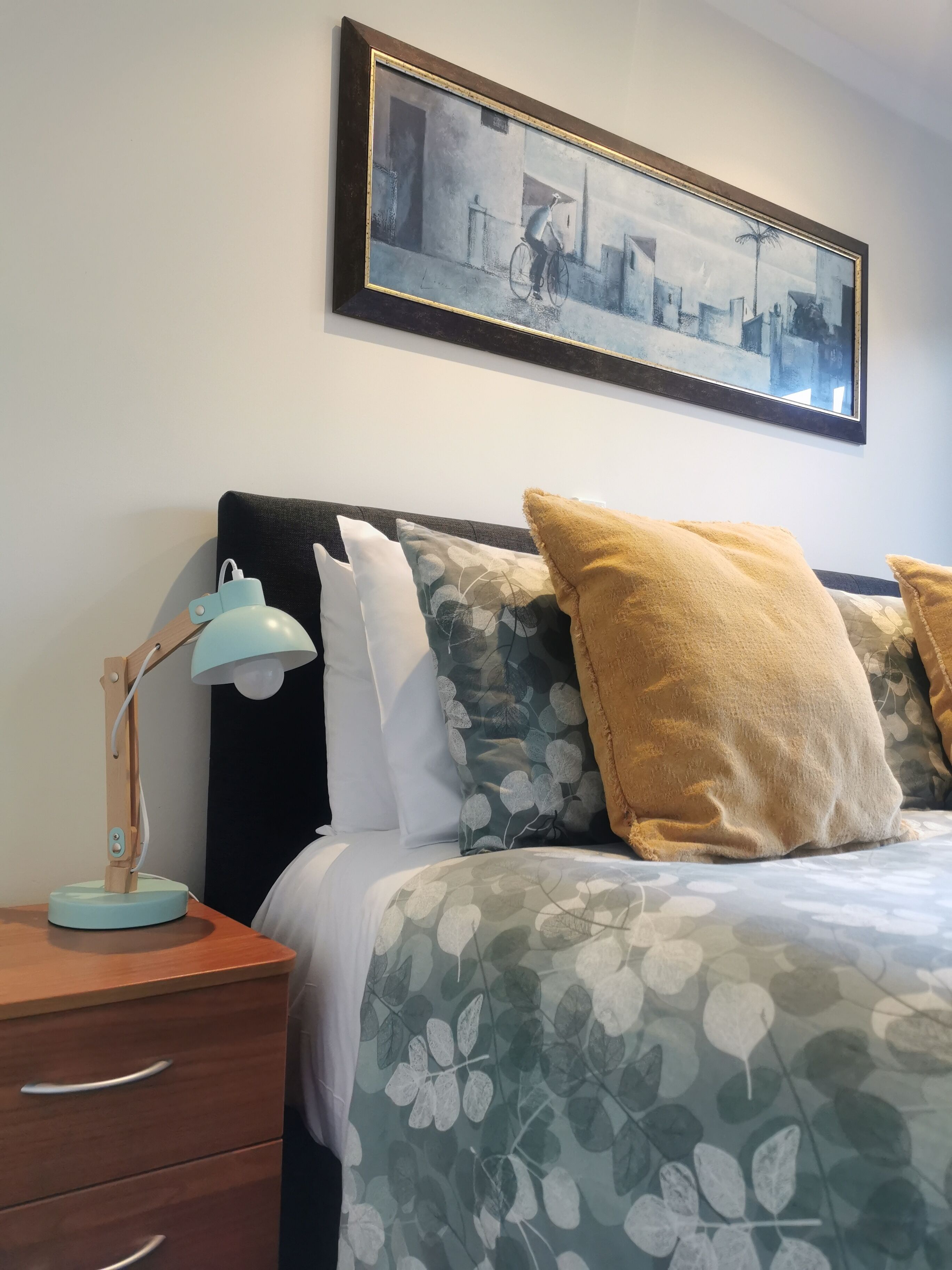 A neatly arranged bedroom with a bed, pillows, a bedside table, a lamp, and a framed artwork on the wall.