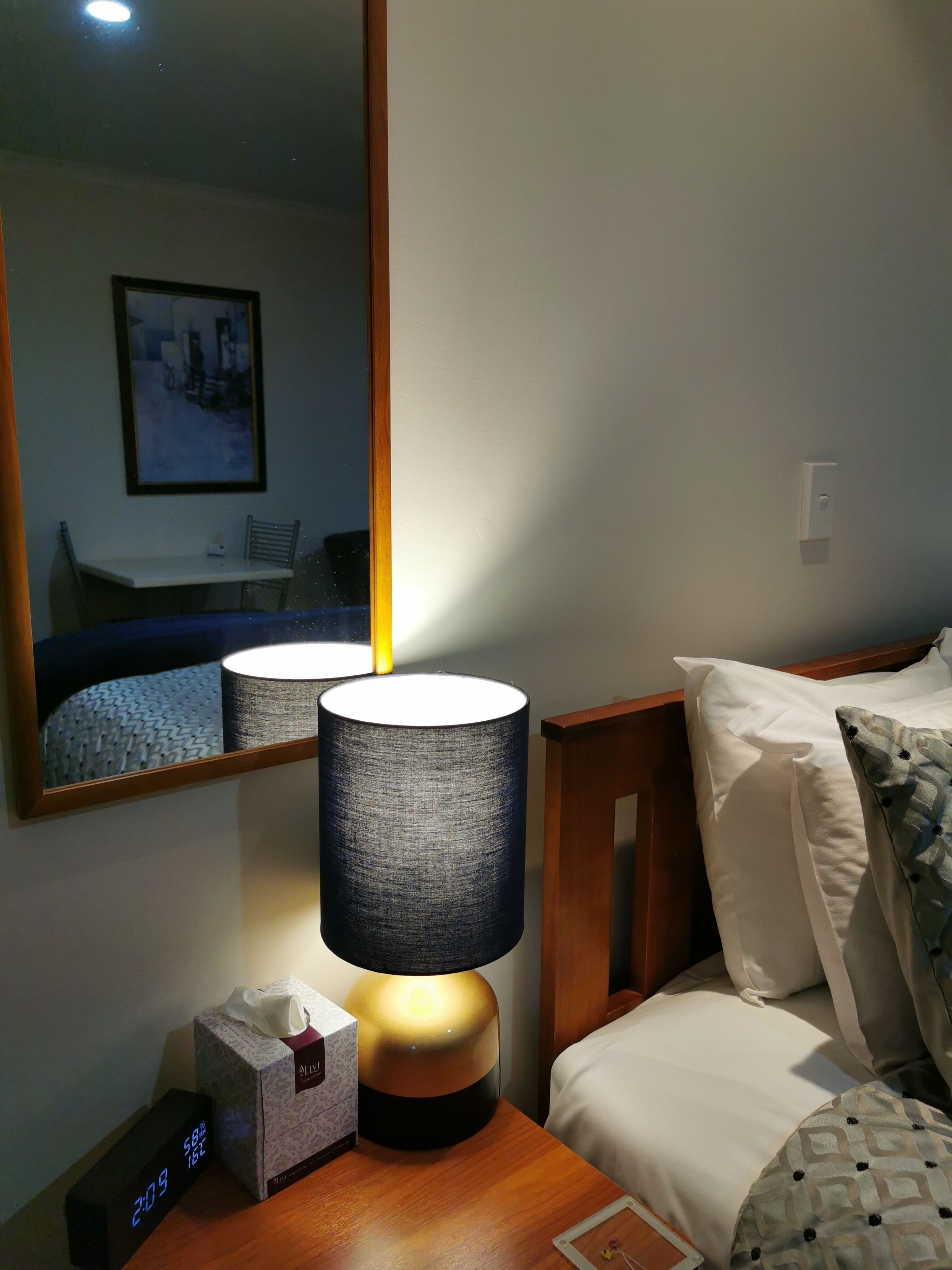A hotel room with a bedside table, a lamp, a digital clock, a tissue box, and a bed with pillows.