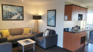 A modern living room with a sofa, armchair, and coffee table, a kitchen with wooden cabinets and a microwave, and two framed artworks on the walls.