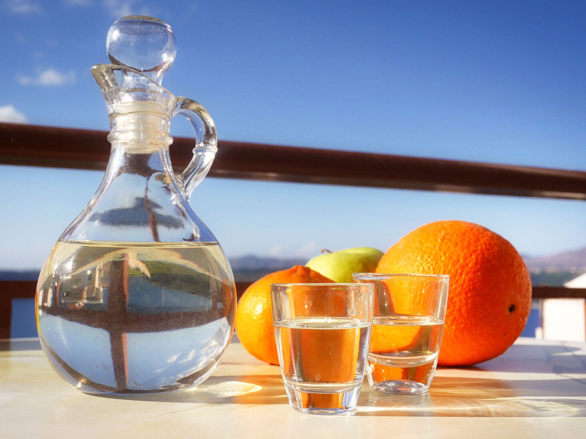 A clear glass pitcher and two shot glasses on a table with an orange and a pear.