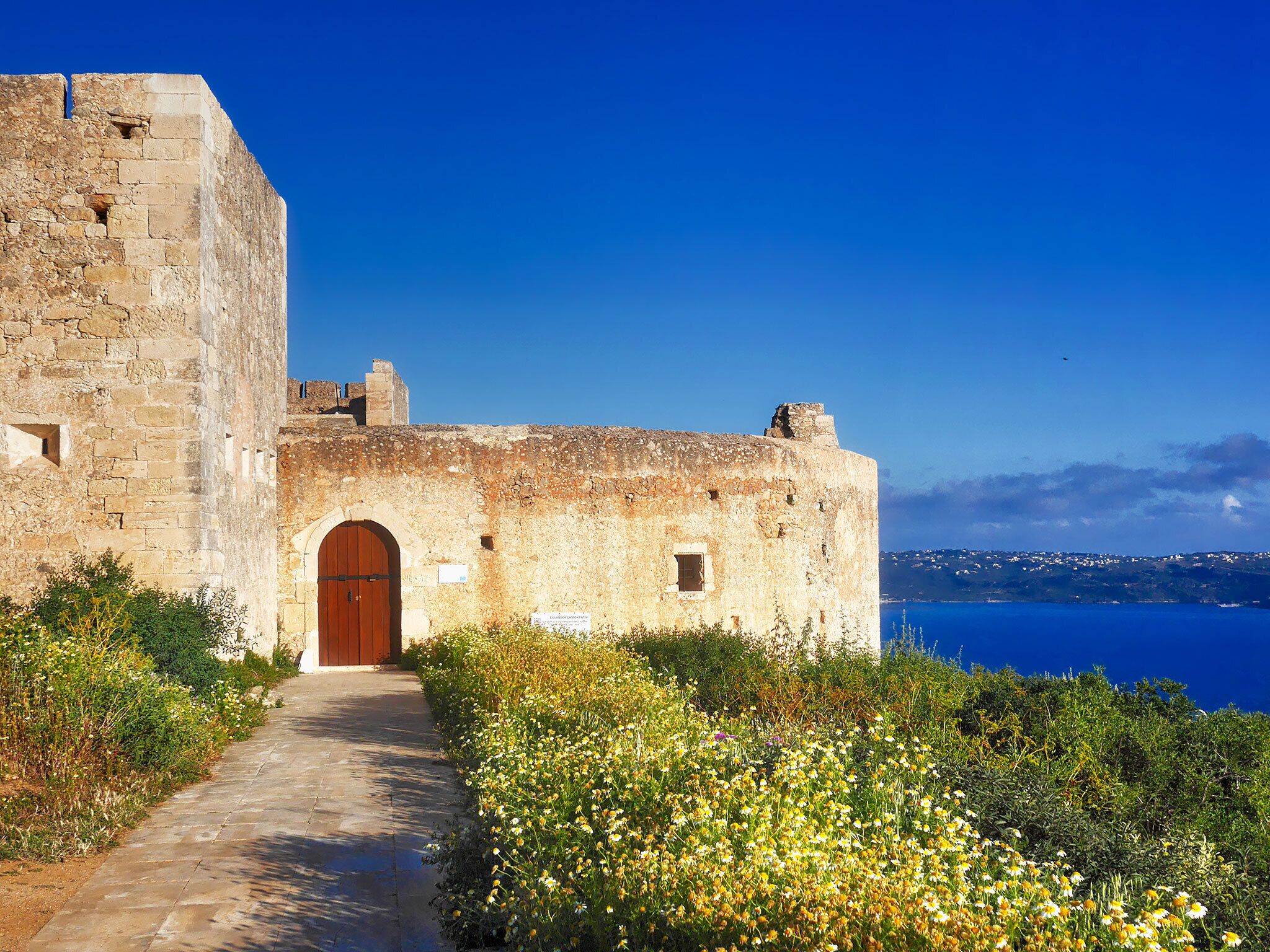 Ein historisches Steinhaus mit einem zentralen Rundbogenportal, einem gepflasterten Weg davor und einem Blick auf das Meer im Hintergrund.