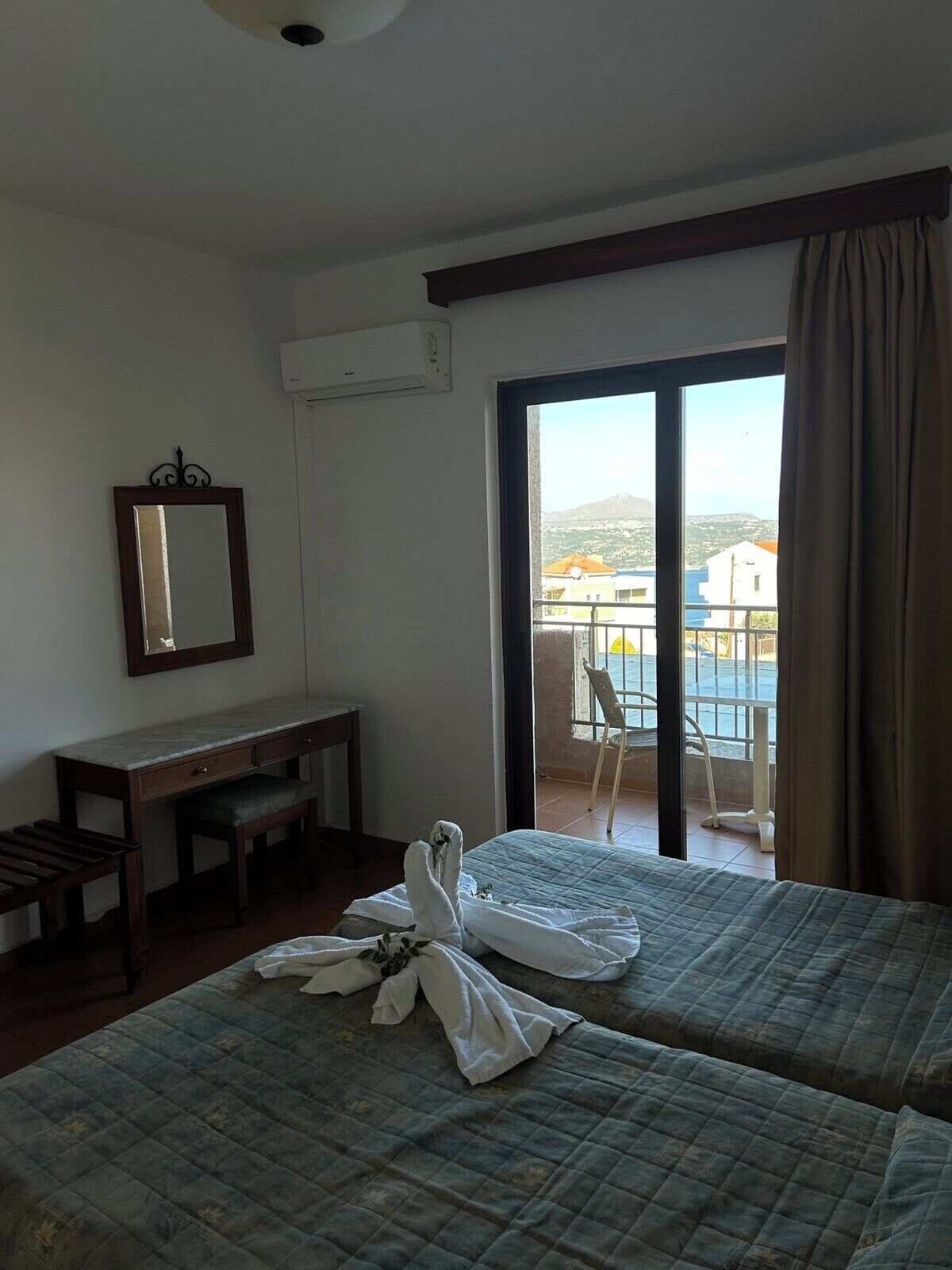 Habitación de hotel con dos camas, un escritorio, una silla y un ventanal con vistas a edificios y montañas.