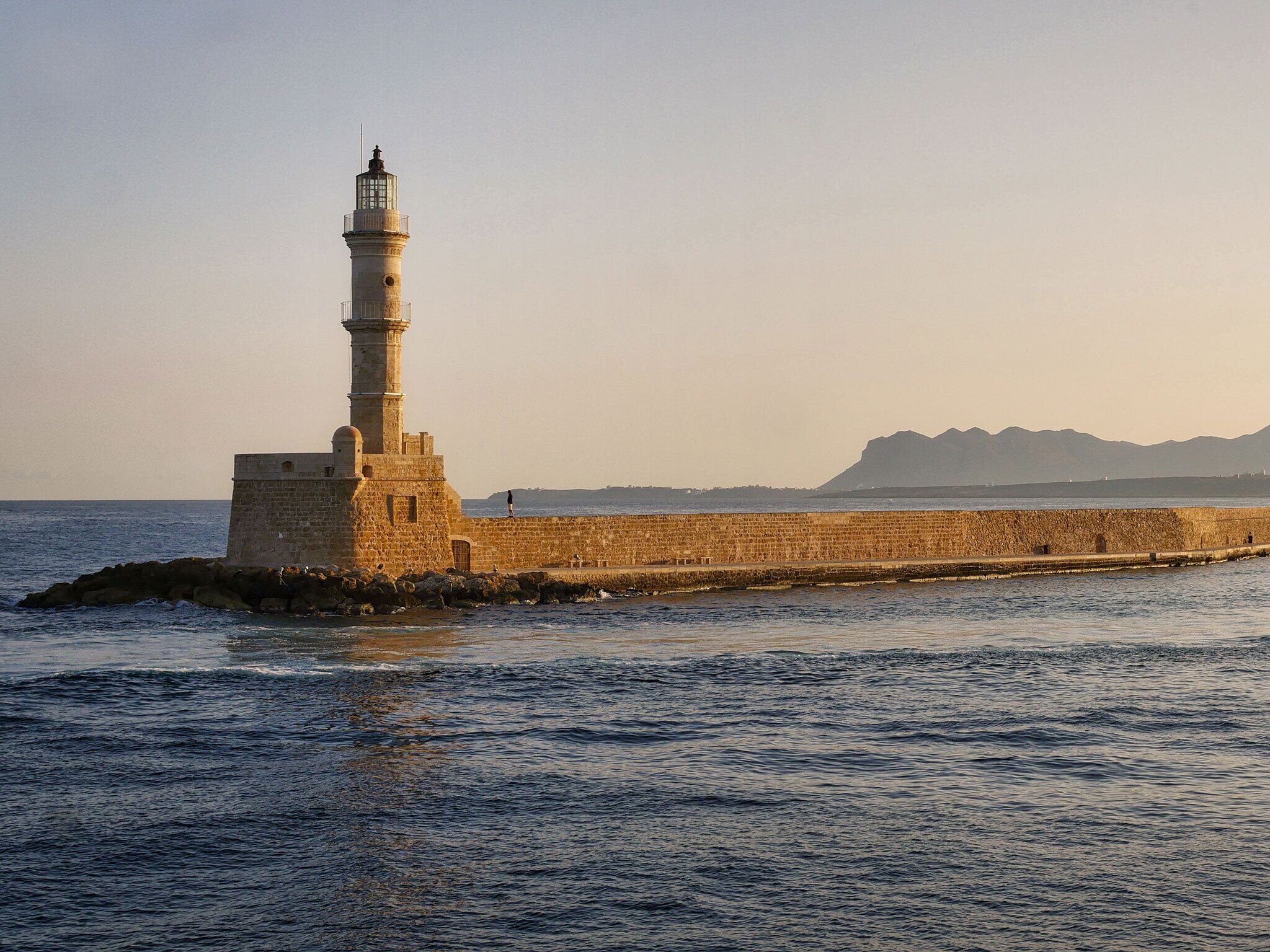 A lighthouse on a rocky outcrop with a clear view of the sea and distant mountains.