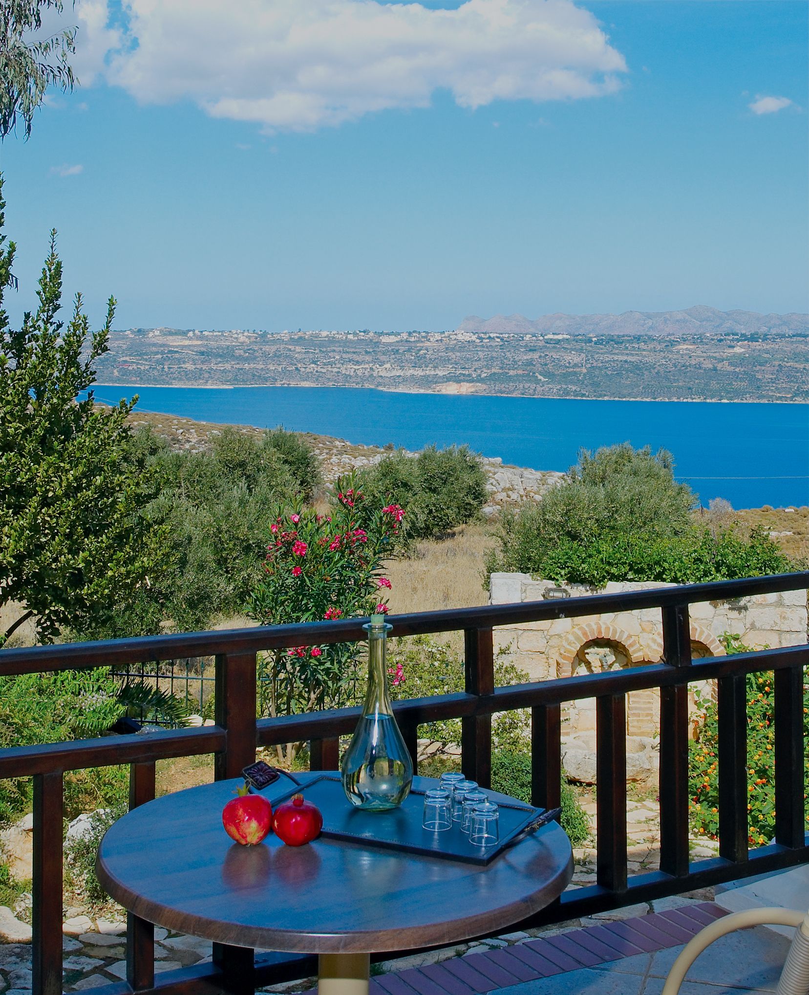 Una mesa redonda con un jarrón, una granada y vasos en un balcón con vista a un paisaje costero, mar azul transparente y colinas lejanas.