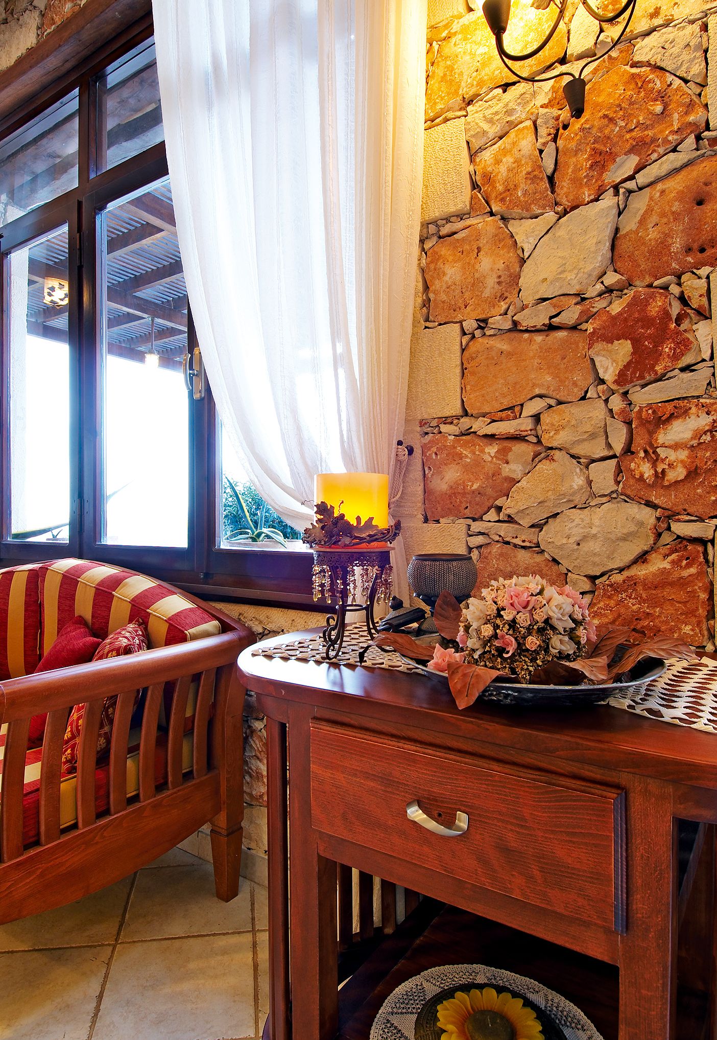 A cozy room with a stone wall, a wooden chair with cushions, a table with a vase of flowers, and a window with curtains.