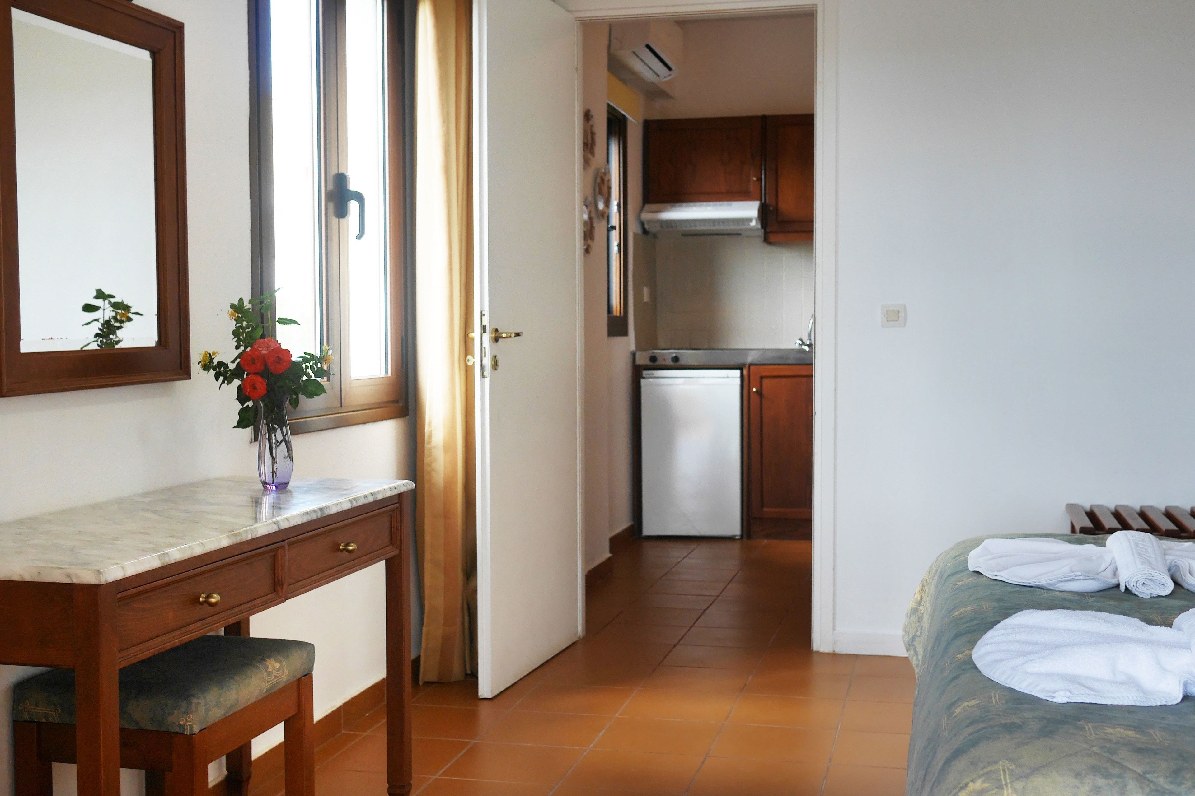 Une chambre d’hôtel comprenant un lit, un bureau avec des tiroirs, une fenêtre avec des rideaux, un coin cuisine avec des placards et un lave-vaisselle.