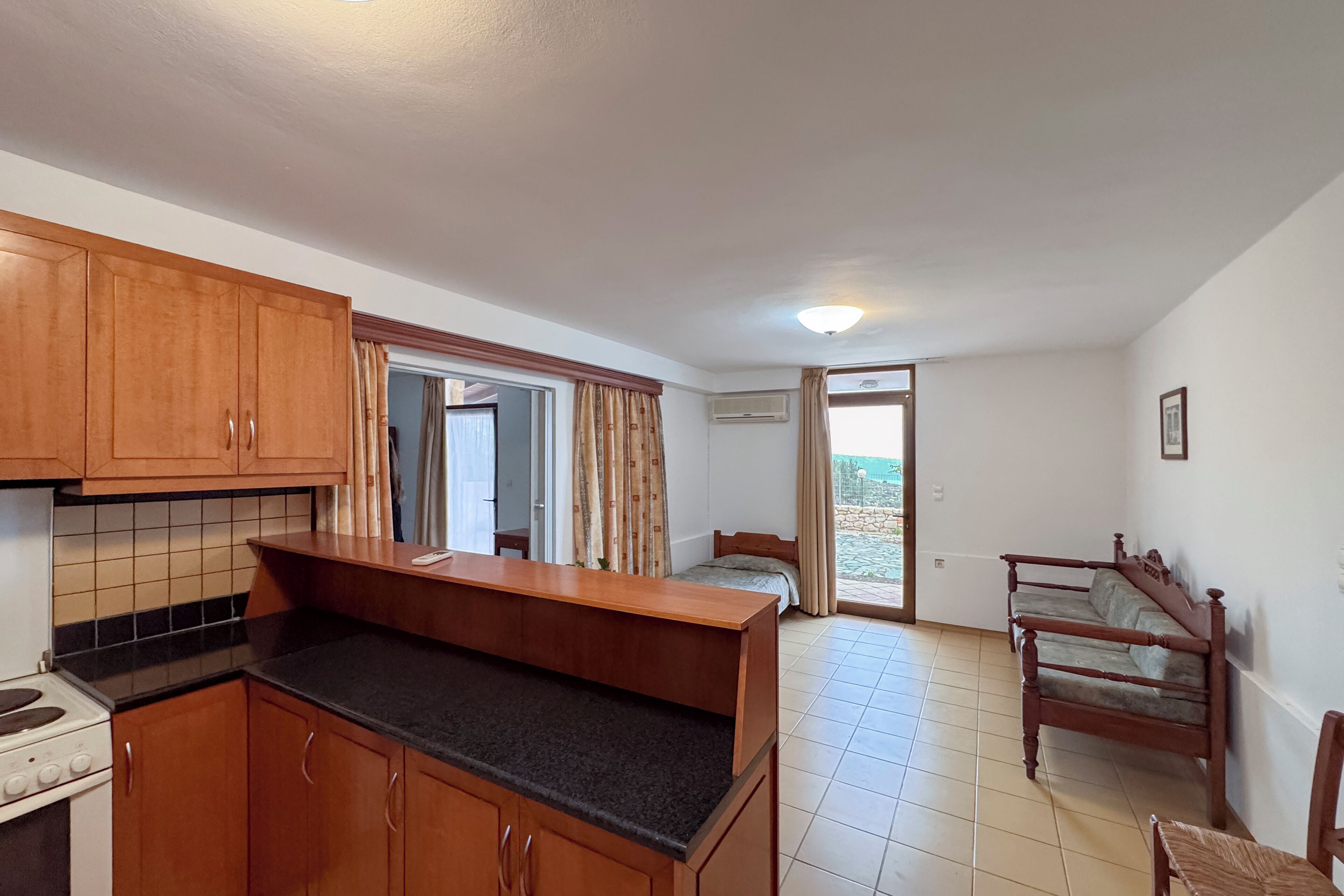 A compact kitchen with wooden cabinets and a countertop, a small dining table, a bed, and a chair in a room with a tiled floor and a door leading outside.