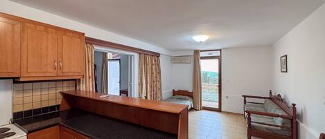 A compact kitchen with wooden cabinets and a countertop, a small dining table, a bed, and a chair in a room with a tiled floor and a door leading outside.